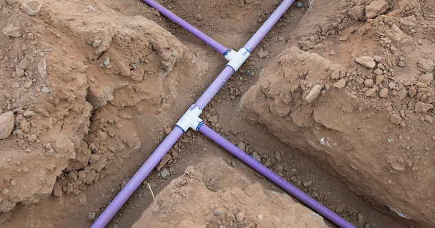 Purple PVC pipes with white fittings are laid out in a cross configuration in an excavated dirt trench, likely part of a plumbing or utility installation project.